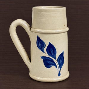 May include: A cylindrical ceramic beer mug with a cream-colored glaze and a single curved handle. The body of the mug features a hand-painted cobalt blue leaf motif consisting of four distinct leaves arranged on a stem.