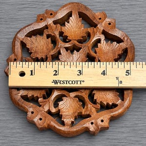 May include: A brown wooden coaster with an intricate openwork design featuring carved maple leaves. A Westcott ruler is placed across the center, showing the coaster measures approximately 5.125 inches in width at its widest point.
