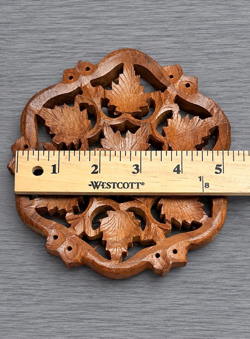 May include: A brown wooden coaster with an intricate openwork design featuring carved maple leaves. A Westcott ruler is placed across the center, showing the coaster measures approximately 5.125 inches in width at its widest point.