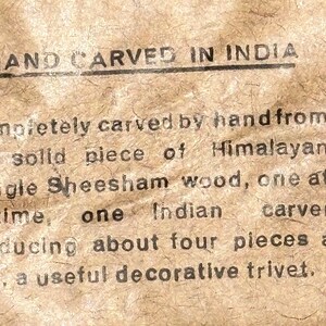 May include: A tan textured paper label with black printed text that reads: HAND CARVED IN INDIA. Completely carved by hand from one solid piece of Himalayan Jungle Sheesham wood, one at a time, one Indian carver producing about four pieces a day, a useful decorative trivet.