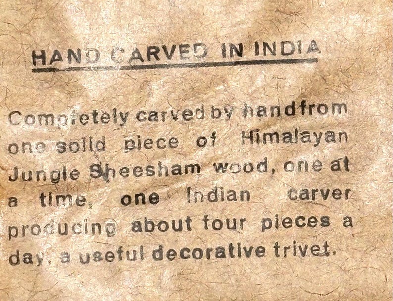 May include: A tan textured paper label with black printed text that reads: HAND CARVED IN INDIA. Completely carved by hand from one solid piece of Himalayan Jungle Sheesham wood, one at a time, one Indian carver producing about four pieces a day, a useful decorative trivet.