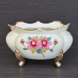 May include: A white porcelain planter with a scalloped rim featuring a gold-painted edge. The body is decorated with a hand-painted floral motif of pink flowers and green leaves. The vessel is supported by three metallic gold-toned feet.