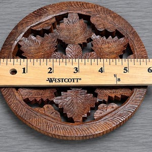 May include: A circular brown wooden coaster featuring an intricate carved leaf pattern. A Westcott ruler is placed across the center, showing the coaster has a diameter of approximately 5.5 inches. The wood has a dark, natural grain finish.