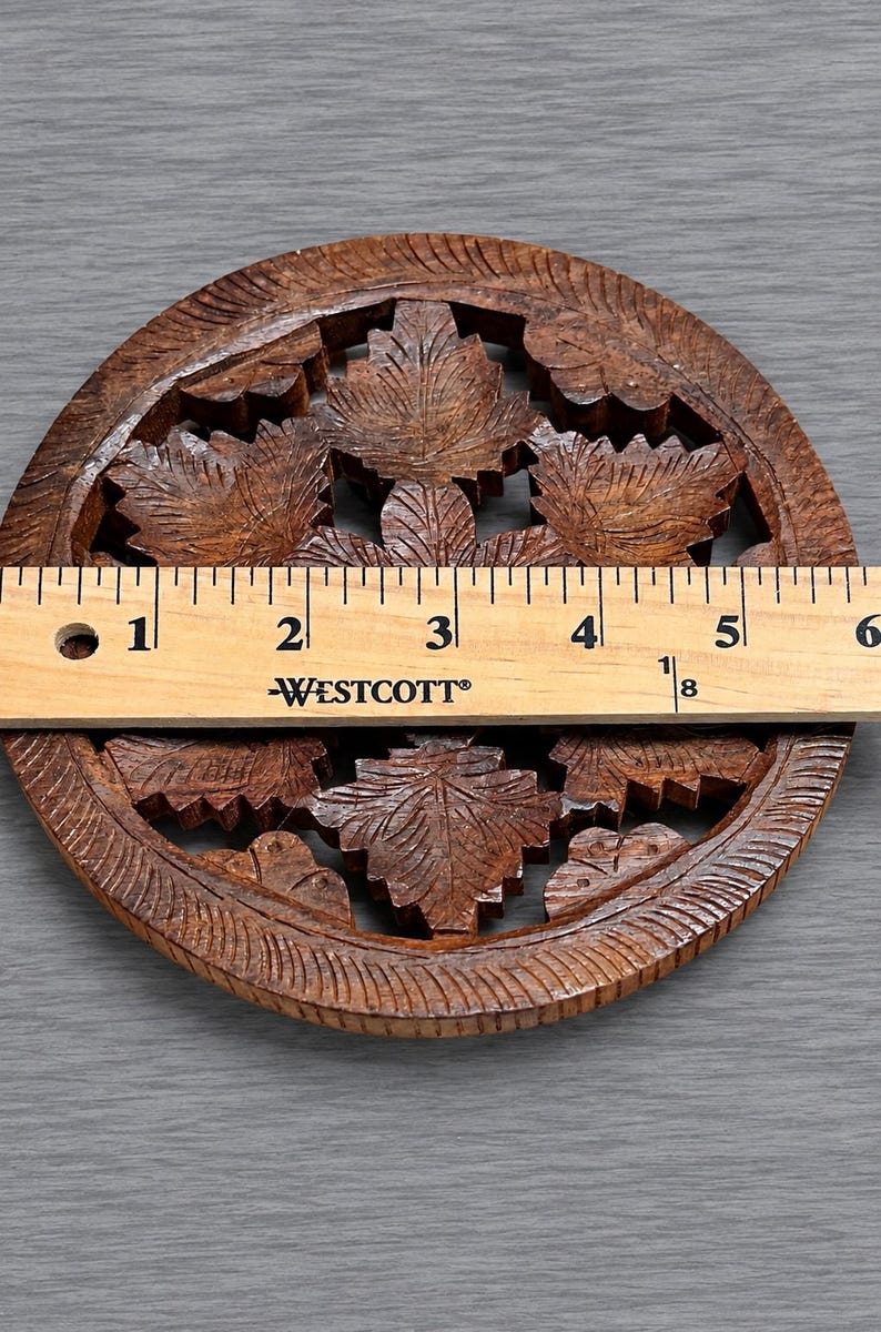 May include: A circular brown wooden coaster featuring an intricate carved leaf pattern. A Westcott ruler is placed across the center, showing the coaster has a diameter of approximately 5.5 inches. The wood has a dark, natural grain finish.