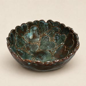 May include: A round ceramic bowl featuring a textured surface covered in embossed heart shapes. The bowl has a dark brown base glaze with a mottled teal and green finish applied over the hearts, creating a deep, layered appearance with a scalloped rim.