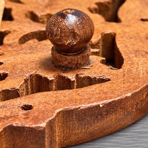 May include: A circular brown wooden board featuring a central spherical knob handle and multiple irregular cut-out shapes throughout the surface. The wood displays a textured, natural grain pattern with visible carved edges and a rustic finish.