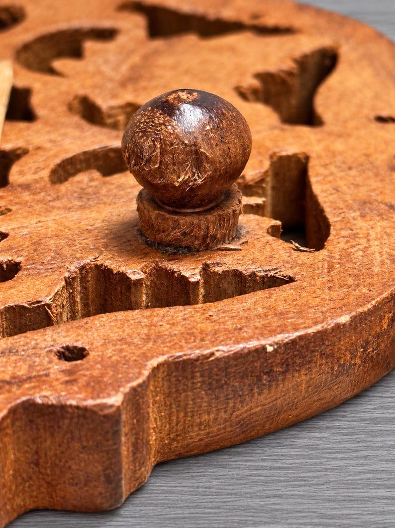 May include: A circular brown wooden board featuring a central spherical knob handle and multiple irregular cut-out shapes throughout the surface. The wood displays a textured, natural grain pattern with visible carved edges and a rustic finish.