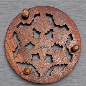 May include: A circular brown wooden trivet featuring an intricate laser-cut floral or snowflake pattern. The piece includes three small spherical wooden feet attached to the underside and the word INDIA is stamped in the center of the design.
