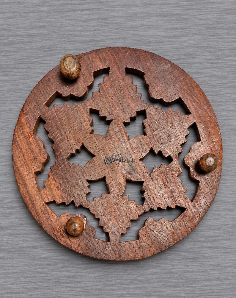 May include: A circular brown wooden trivet featuring an intricate laser-cut floral or snowflake pattern. The piece includes three small spherical wooden feet attached to the underside and the word INDIA is stamped in the center of the design.