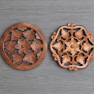 May include: Two circular and square-shaped wooden trivets featuring intricate, hand-carved floral and leaf patterns. The natural brown wood grain is visible throughout both pieces, which display symmetrical cut-out designs and a smooth, polished finish.