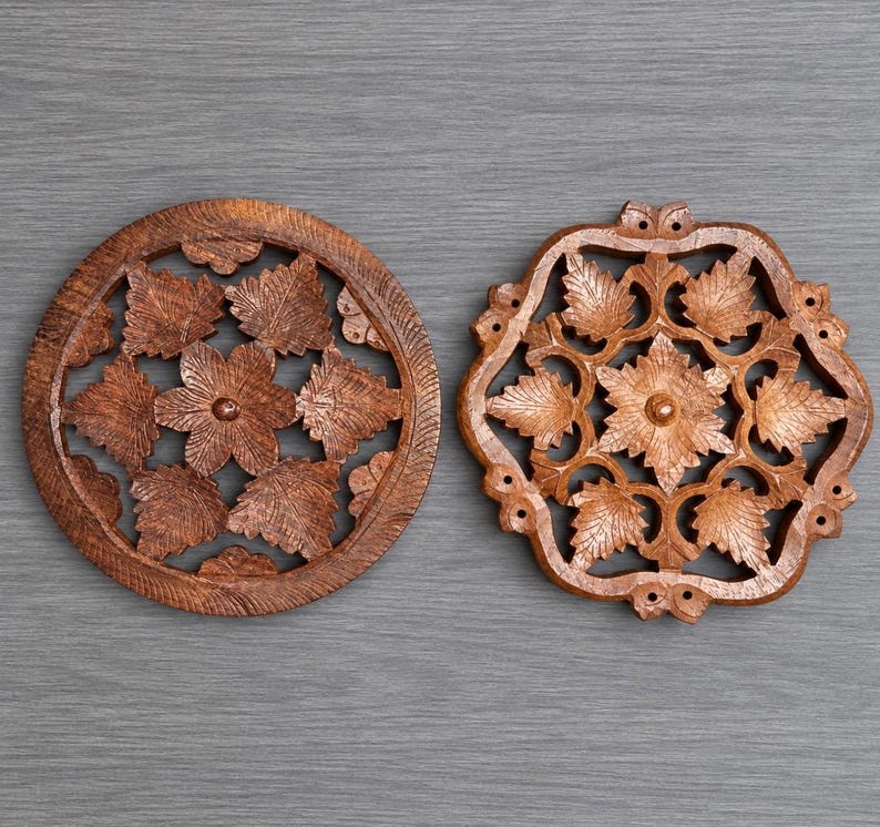May include: Two circular and square-shaped wooden trivets featuring intricate, hand-carved floral and leaf patterns. The natural brown wood grain is visible throughout both pieces, which display symmetrical cut-out designs and a smooth, polished finish.