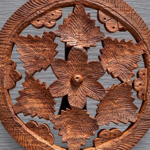 May include: A circular brown wooden trivet featuring an intricate openwork design. The center displays a six-petaled flower surrounded by detailed leaf motifs. The outer ring is textured with carved lines, and the entire piece has a natural wood grain.