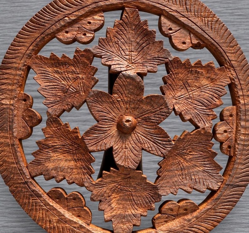 May include: A circular brown wooden trivet featuring an intricate openwork design. The center displays a six-petaled flower surrounded by detailed leaf motifs. The outer ring is textured with carved lines, and the entire piece has a natural wood grain.