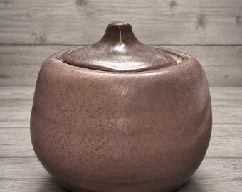 May include: A round, hand-thrown ceramic sugar bowl with a dusty mauve, speckled glaze. The vessel features a bulbous body and a matching lid with a small, pointed central handle. The surface has a matte, textured finish with subtle color variations.