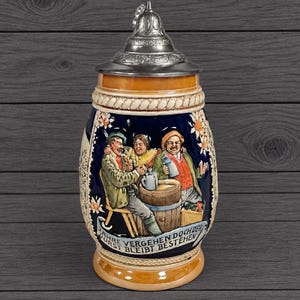May include: A ceramic beer stein with a metal lid featuring a relief scene of three people drinking around a barrel. The text on the base reads JAHRE VERGEHEN DOCH DER DURST BLEIBT BESTEHEN. The stein has a blue background with floral accents.