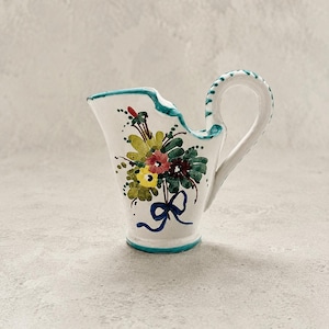 May include: A white ceramic creamer pitcher featuring a hand-painted floral bouquet in shades of yellow, pink, and green, accented with a blue ribbon motif. The pitcher has a teal rim, a curved handle with teal detailing, and a flared spout.