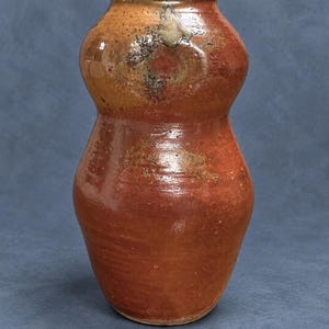 May include: A gourd-shaped ceramic vase with a glossy, mottled finish in shades of deep rust, burnt orange, and earthy brown. The surface features subtle variations in texture and color, with a smooth, rounded silhouette and a narrow neck opening.
