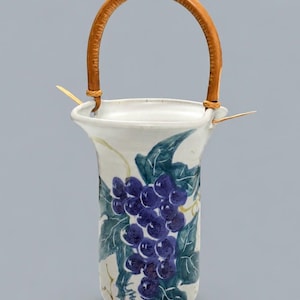 May include: A white ceramic vase with a flared rim and a curved bamboo handle attached by two wooden pins. The exterior features a hand-painted design of a cluster of purple grapes with green leaves and thin, winding brown vines against the white surface.