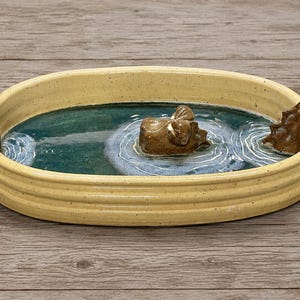 May include: An oval ceramic dish with a speckled cream-colored rim and a deep teal interior basin. The center features a brown sculpted hippopotamus partially submerged in water, with a small white figure on one side and a brown rock on the other.
