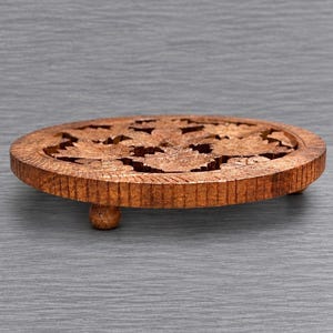 May include: A round, brown wooden trivet featuring an intricate, carved floral pattern across its surface. The circular piece is supported by three small, rounded feet, and the outer edge displays a textured, vertical grooved design.