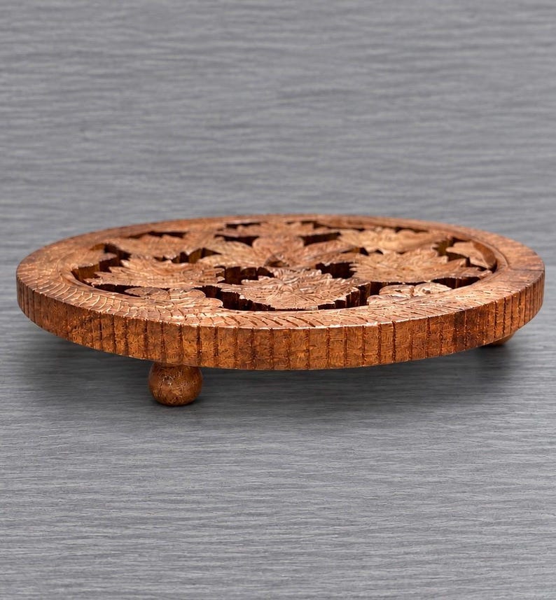 May include: A round, brown wooden trivet featuring an intricate, carved floral pattern across its surface. The circular piece is supported by three small, rounded feet, and the outer edge displays a textured, vertical grooved design.