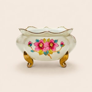 May include: A cream-colored porcelain bowl with a scalloped rim and three gold-toned feet. The exterior features a hand-painted floral design with vibrant pink flowers, green leaves, and blue accents, finished with a delicate gold-painted edge.