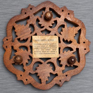 May include: A brown wooden trivet with an intricate, hand-carved openwork leaf pattern. The center features a square paper label with the text: HAND CARVED IN INDIA. Completely carved by hand from one solid piece of Himalayan Jungie Sheesham wood.