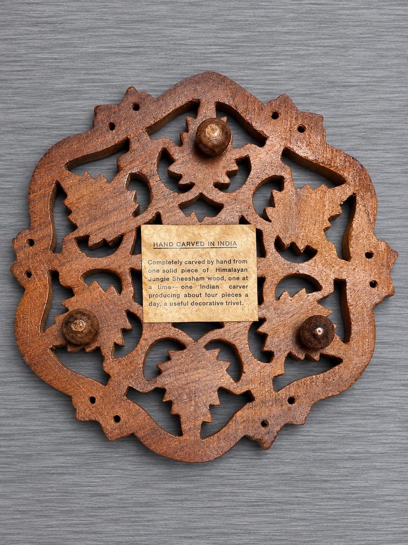 May include: A brown wooden trivet with an intricate, hand-carved openwork leaf pattern. The center features a square paper label with the text: HAND CARVED IN INDIA. Completely carved by hand from one solid piece of Himalayan Jungie Sheesham wood.