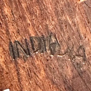 May include: A close-up view of a textured, brown wooden surface featuring the word INDIA stamped or burned into the grain. The wood displays a natural, fibrous pattern with vertical striations and dark brown tones throughout the material.
