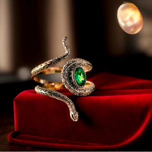 May include: A gold snake bracelet with a green gemstone set in a diamond-like setting. The bracelet is on a red velvet surface.