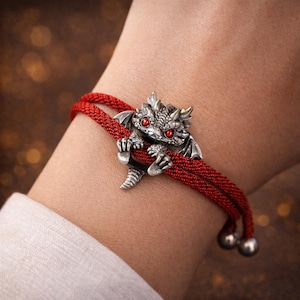 Puede incluir: Una pulsera con un dije de dragón plateado y un cordón rojo. El dragón tiene ojos rojos y se aferra al cordón. La pulsera tiene dos cuentas plateadas al final del cordón.