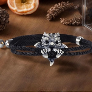 Könnte beinhalten: Ein schwarzes Kordelarmband mit einem silberfarbenen Drachenanhänger in der Mitte. Der Drache hat detaillierte Schuppen, kleine Flügel und dunkle Augen, wobei seine Krallen die doppelte schwarze Kordel umgreifen. Das Armband hat silberfarbene Endkappen.