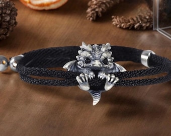 May include: A black cord bracelet featuring a central silver-toned dragon charm. The dragon has detailed scales, small wings, and dark eyes, with its claws gripping the double-stranded black cord. The bracelet includes silver-toned metal end caps.