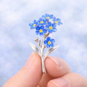 Könnte beinhalten: Silberfarbene Brosche in Form einer Vergissmeinnicht-Blume. Die Brosche zeigt mehrere blaue Blüten mit gelben Zentren und grünen Blättern. Die Brosche wird in der Hand einer Person gehalten.