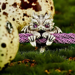 May include: A silver-toned dragon bead featuring intricate scale detailing and two deep magenta gemstone eyes. The dragon is perched on a textured purple cord, with its front claws gripping the material and its tail extending downward.