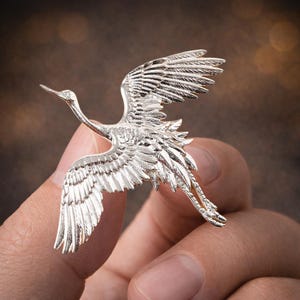 May include: A detailed silver-colored crane brooch in mid-flight. The bird's wings are outstretched, showcasing intricate feather detailing. The brooch is held in a hand, with a blurred background.