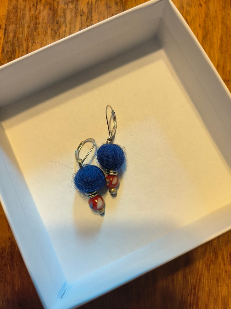 May include: A pair of blue wool ball earrings with red and white bead accents. The earrings are silver and have a hook closure.