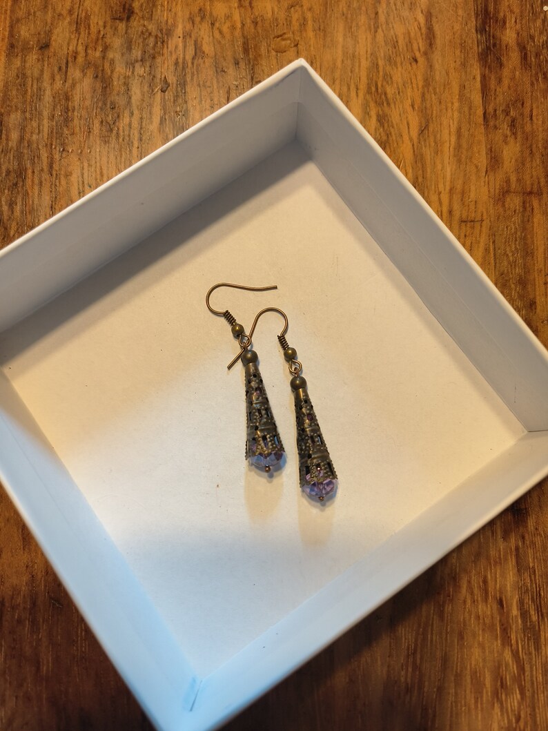 May include: A pair of dangle earrings with a purple glass bead at the bottom and a gold filigree design. The earrings are in a white box.