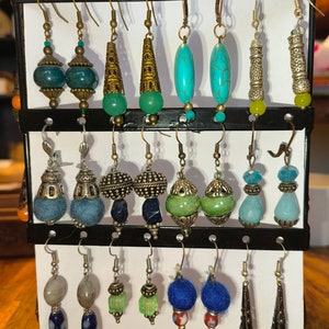 May include: A display of 12 pairs of earrings with various styles and colors, including turquoise, blue, green, and silver. The earrings are displayed on a white background with a black metal rack.