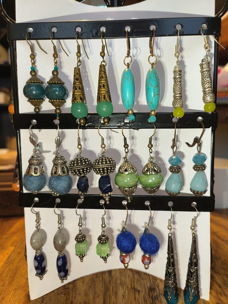 May include: A display of 12 pairs of earrings with various styles and colors, including turquoise, blue, green, and silver. The earrings are displayed on a white background with a black metal rack.
