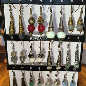 May include: A display of 18 pairs of dangle earrings with various colored beads, stones, and metal accents. The earrings are arranged on a black stand with hooks.