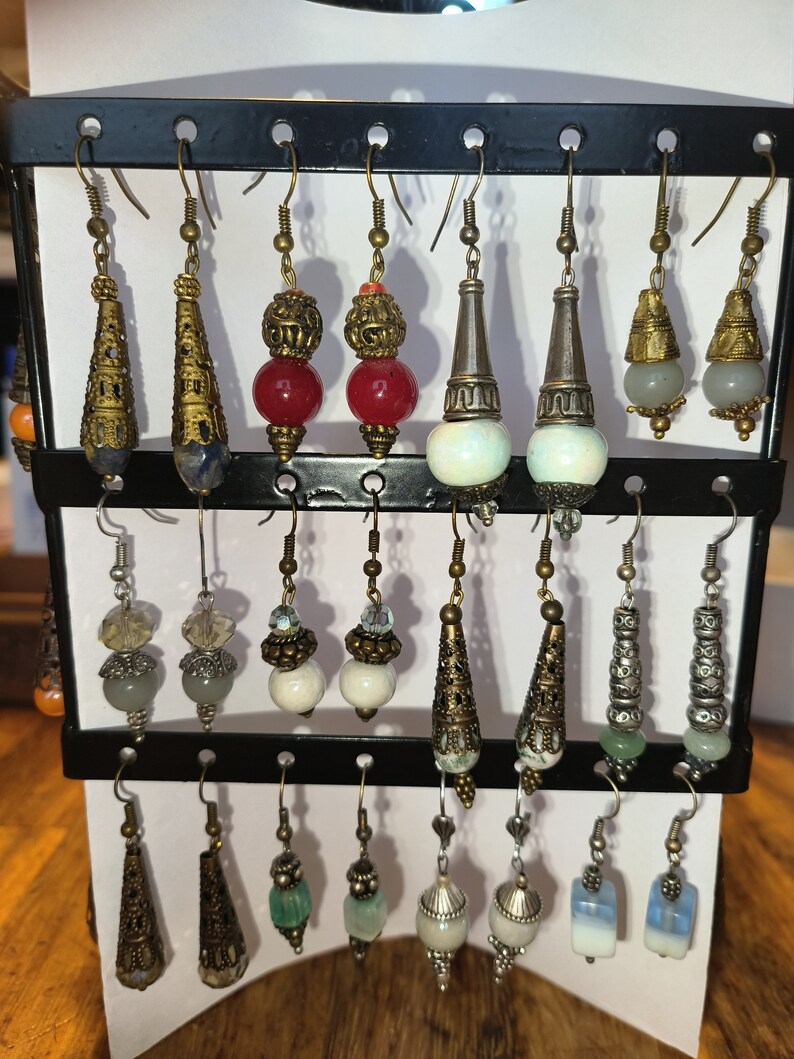 May include: A display of 18 pairs of dangle earrings with various colored beads, stones, and metal accents. The earrings are arranged on a black stand with hooks.