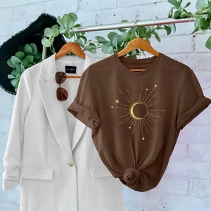 Op de afbeelding: Een bruin t-shirt met een gouden halve maan en zon grafische afbeelding. De afbeelding heeft een ster en kleine stippen eromheen. Het t-shirt hangt aan een houten hanger.