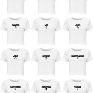 May include: A collection of twelve white short-sleeved t-shirts arranged in a grid. Each shirt features a black graphic print of a zodiac sign name and its corresponding astrological symbol, including Aries, Taurus, Gemini, Cancer, Leo, Virgo, Libra, Scorpio, Sagittarius, Capricorn, Aquarius, and Pisces.