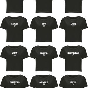 May include: A collection of twelve black short-sleeved crop tops, each featuring a unique white graphic print of a zodiac sign name and its corresponding astrological symbol, including Aries, Taurus, Gemini, Cancer, Leo, Virgo, Libra, Scorpio, Sagittarius, Capricorn, Aquarius, and Pisces.