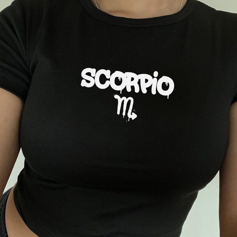 May include: A black short-sleeved t-shirt featuring the word SCORPIO in white graffiti-style lettering across the chest, positioned above the white Scorpio zodiac symbol. The design has a dripping paint effect on the letters and the symbol.