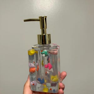May include: Clear, rectangular soap dispenser with a gold-colored pump. The dispenser is filled with colorful, miniature duck figurines. The ducks are various colors, including pink, blue, yellow, and green. The dispenser is held in a hand.