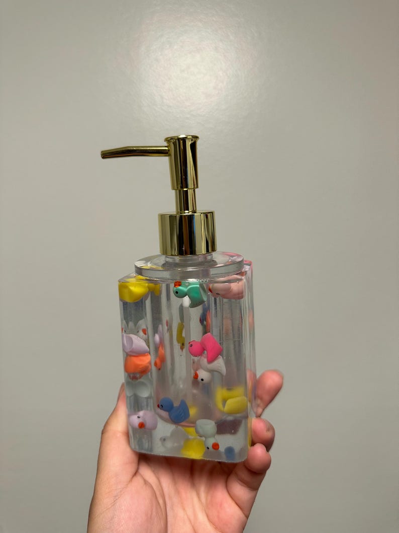 May include: Clear, rectangular soap dispenser with a gold-colored pump. The dispenser is filled with colorful, miniature duck figurines. The ducks are various colors, including pink, blue, yellow, and green. The dispenser is held in a hand.