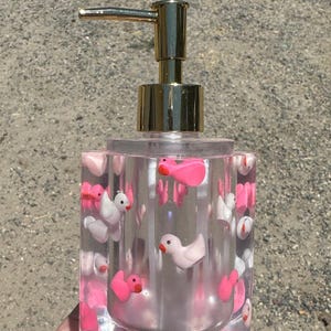 May include: Clear soap dispenser with a gold pump, filled with pink and white duck-shaped embellishments. The dispenser has a square shape and is made of clear resin, creating a whimsical and decorative bathroom accessory.