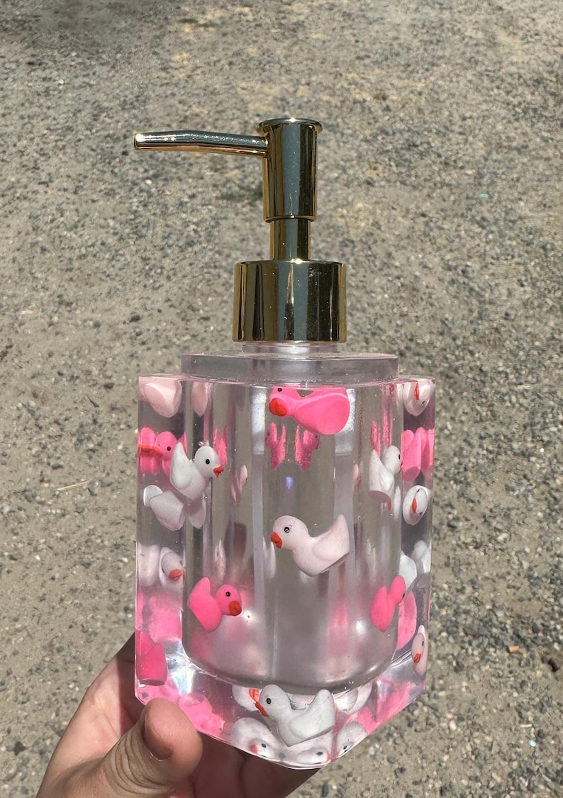 May include: Clear soap dispenser with a gold pump, filled with pink and white duck-shaped embellishments. The dispenser has a square shape and is made of clear resin, creating a whimsical and decorative bathroom accessory.